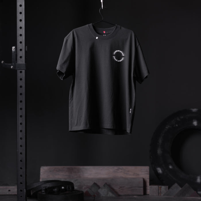Black Tech Essential Relaxed Tee with circular logo, displayed on hanger in gym setting