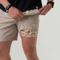 Close-up of a man wearing khaki athletic shorts with a camo compression liner underneath. The outer shorts feature a small white logo on the left thigh.