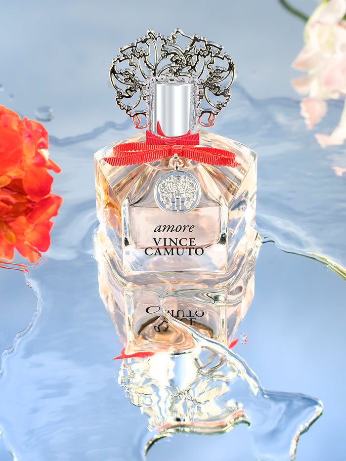 A bottle of Amore 3.4 oz Eau de Parfum Spray by Vince Camuto, adorned with an intricate silver cap and a vibrant red ribbon, rests on a reflective surface amid vivid orange blossoms and shimmering water. The setting reflects the fragrance’s harmonious notes of sparkling fruits and sensual sandalwood, capturing an atmosphere of elegance and allure.