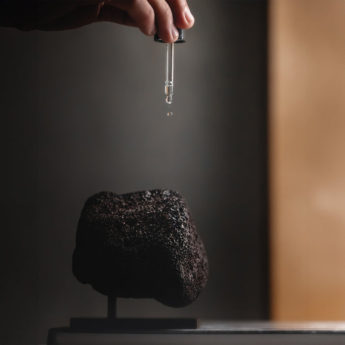 Hand holding dropper above dark textured Essential Oil bottle, demonstrating product application