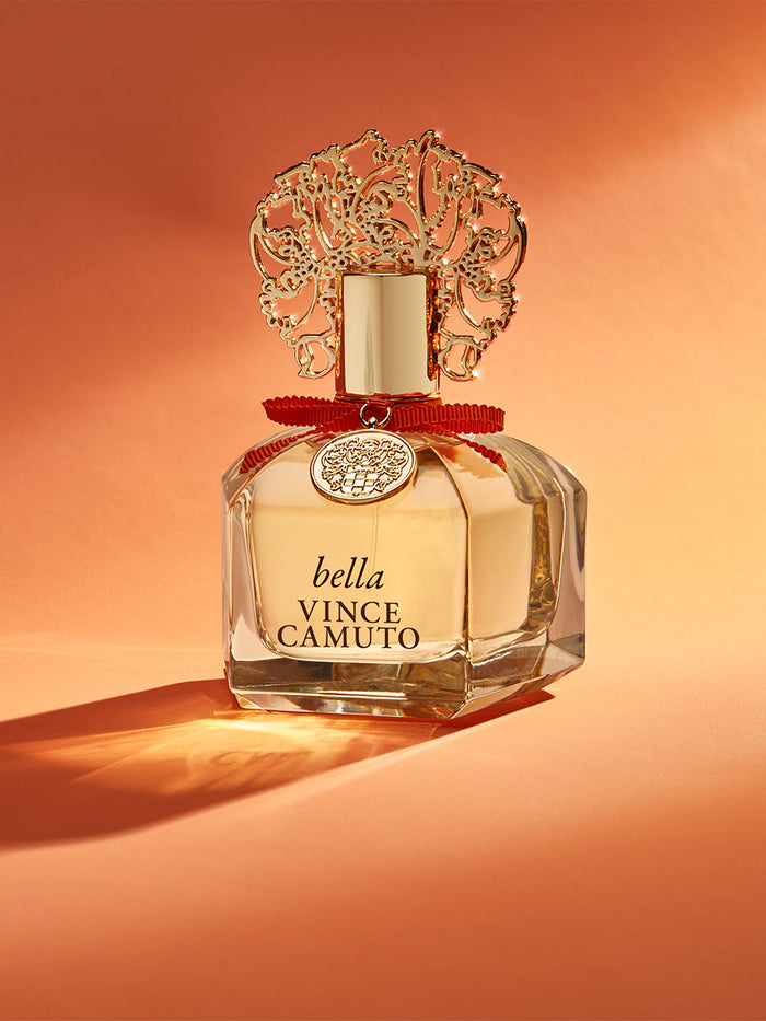 A 1.0 oz bottle of Bella Eau de Parfum Spray by Vince Camuto, featuring a gold ornate cap, red ribbon, and decorative medallion, is displayed against an orange background with a soft shadow. This fragrance blends the bright notes of Italian bergamot and nectarine blossom with the warmth of white amber.