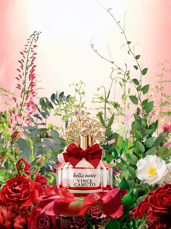 The Bella Notte 3-Piece Set by Vince Camuto is elegantly showcased with a striking red bow, surrounded by vibrant red and white roses and lush green foliage, all set against a gradient pink backdrop—creating a sophisticated scene that embodies the captivating floral essence of this luxurious fragrance set.