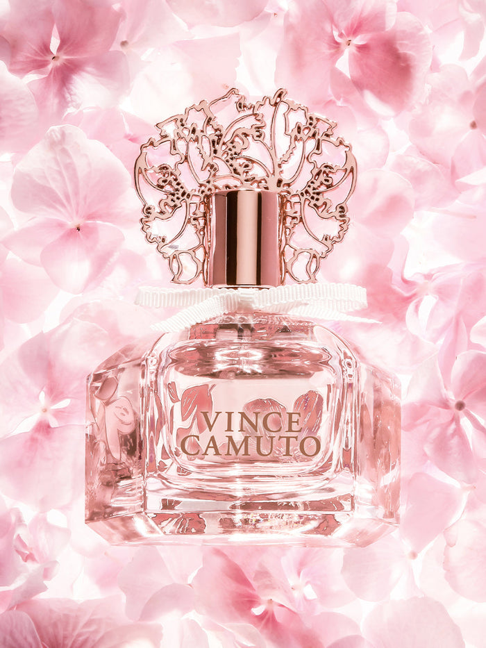 The Brilliante 3.4 oz Eau de Parfum Spray by Vince Camuto features a blush pink bottle with an ornate rose gold cap and delicate white ribbon, surrounded by soft pink flowers. This enchanting fragrance captures the essence of a sparkling scent with elegant white florals, making it an irresistible choice for women.