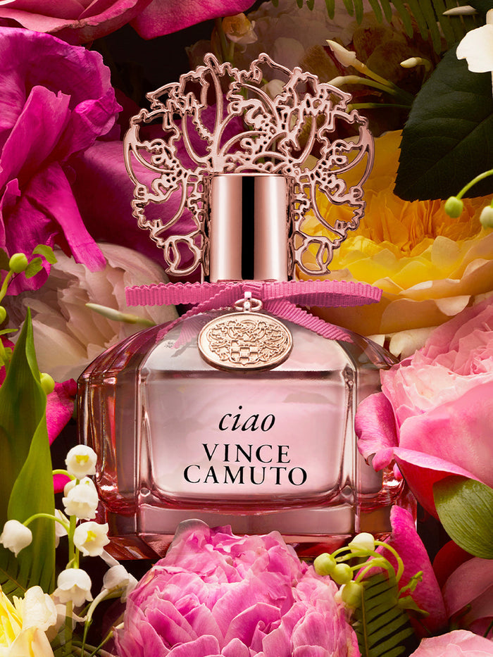 A 3.4 oz bottle of Ciao Eau de Parfum Spray by Vince Camuto, featuring a decorative lid, is surrounded by vibrant pink, yellow, and white flowers, evoking the essence of Mediterranean sea breezes with its fresh citrus notes.