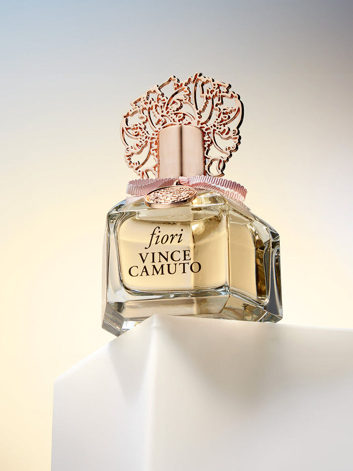 The Fiori 0.2 oz Eau de Parfum Rollerball by Vince Camuto rests on a white pedestal with a soft gradient background, its elegant allure heightened by subtle sandalwood notes and a chic rose gold floral cap.