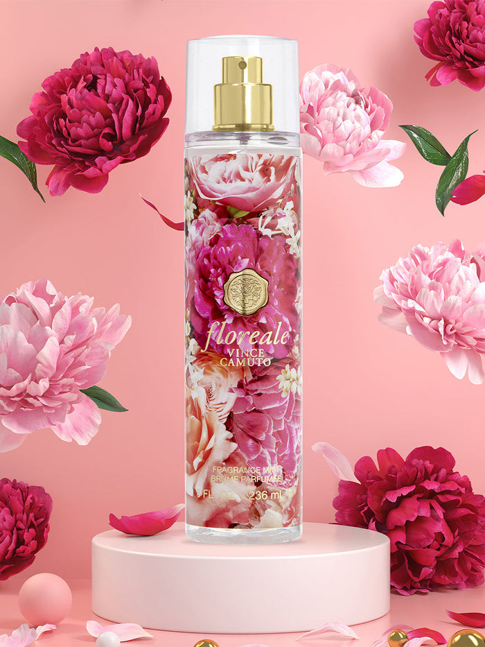 The Vince Camuto Floreale Body Mist 8.0 oz, adorned with pink and white peony motifs, rests elegantly on a round pedestal. Around it, blooming pink and red peonies mingle against a soft pink backdrop, as delicate notes of vanilla and orchid fill the air with an inviting sweetness.