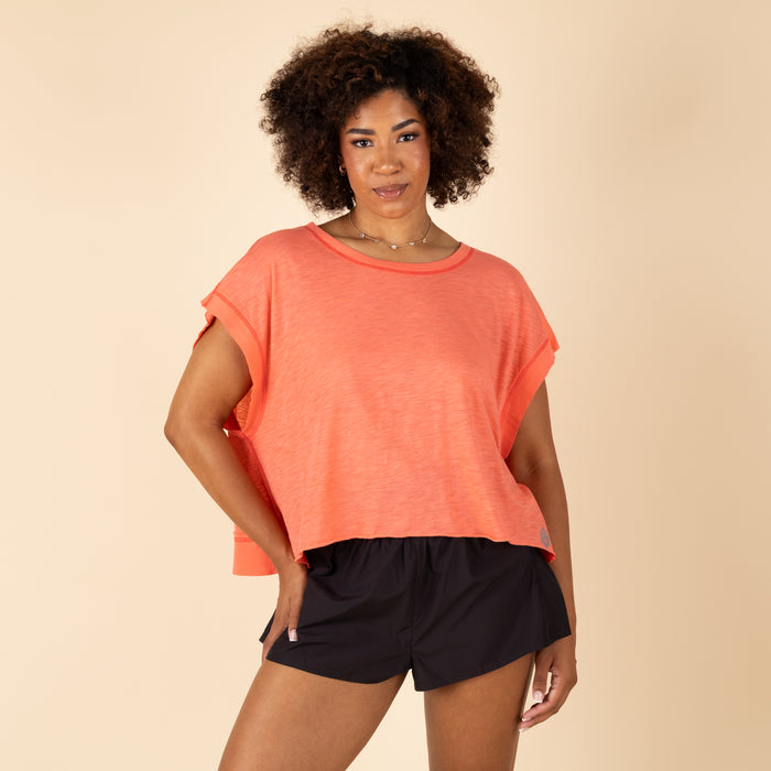 Free People Movement My Time Tee in Neon Coral, sizes XS–XL. Full front view of model wearing the relaxed-fit tee, hands in shorts pockets, showcasing the draped silhouette.