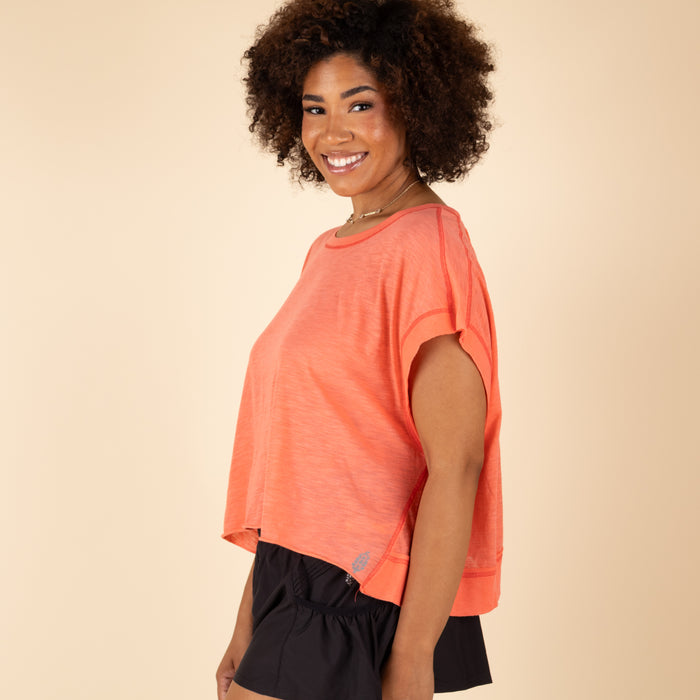 Free People Movement My Time Tee in Neon Coral, sizes XS–XL. Side view of model smiling, highlighting the flowy cut, dropped shoulders, and visible stitching detail.