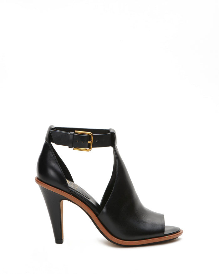 The Vince Camuto Frasper Shootie is a black leather high-heeled peep-toe sandal with an open toe, cutout design, and gold buckle ankle strap, featuring a cushioned footbed for added comfort, shown in side profile on a white background.