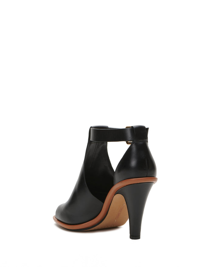 The Vince Camuto Frasper Shootie is a black high-heeled shoe with a closed back, ankle strap, and brown sole featuring a cushioned footbed, shown from the rear on a white background.