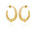 A pair of Vince Camuto Goldtone Open Knotted Hoop Earrings, featuring a chunky, C-shaped hoop with a twisted open knotted design, are displayed upright on a white background with a subtle reflection below, highlighting their bold contemporary style.