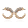 A pair of Vince Camuto Goldtone Pave Crystal Stone Open Hoop Earrings, each featuring one half adorned with shimmering pave crystals and the other half smooth and polished. The earrings are displayed facing each other on a white background.