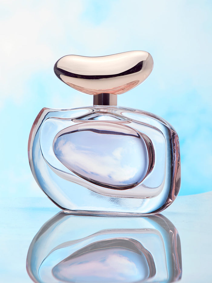 A contemporary, abstract perfume bottle with a rose gold cap—Vince Camuto’s Illuminare 3.4 oz Eau de Parfum—rests on a reflective surface against a soft blue and white sky background, capturing the elegant allure of plum blossom fragrance.