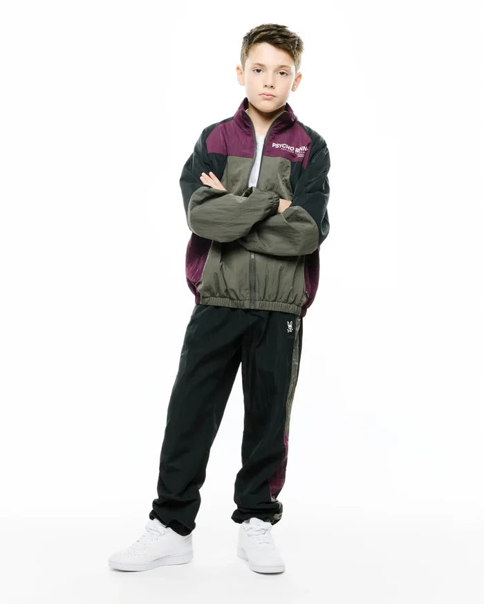 KIDS ALDER NYLON COLORBLOCK PANT in dark green/black with plum side stripe, featuring a small embroidered white Psycho Bunny skull logo on the left thigh. Relaxed fit crinkled nylon, part of a matching colorblock tracksuit.