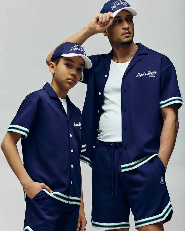 Two people pose confidently in matching Psycho Bunny KIDS BASIL TAPED SHORT - KB0900003, styled with navy baseball shirts, white undershirts, and navy caps against a neutral backdrop.