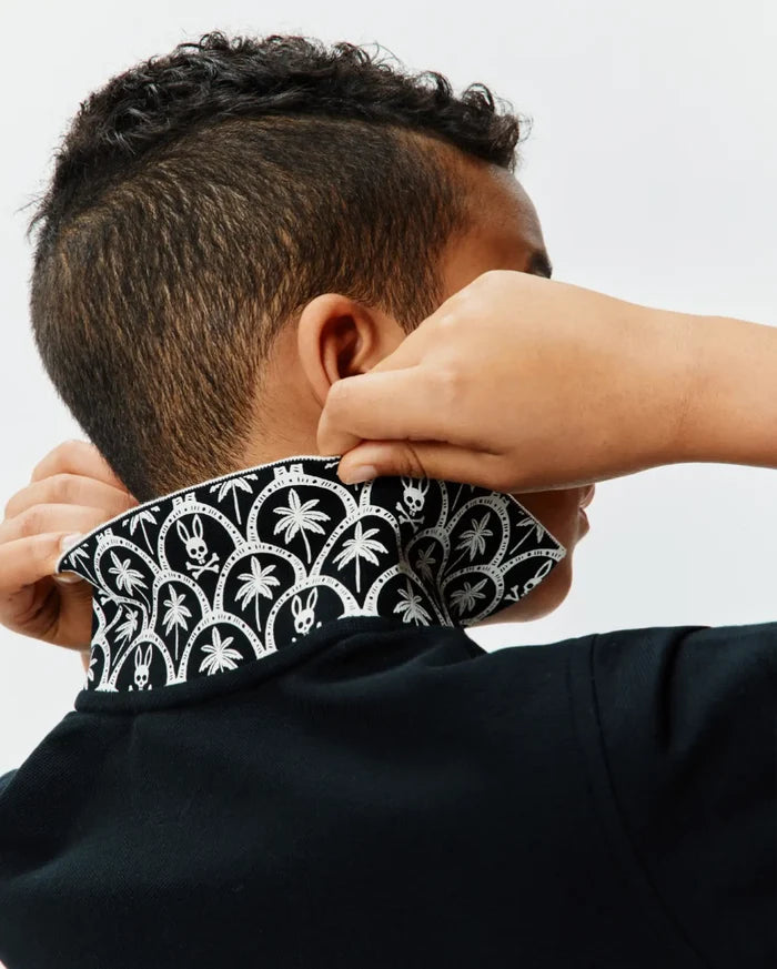 Kids Brad Pique Polo in classic black, featuring a textured pique fabric and a bold, contrasting white pattern on the collar with repeating palm trees and iconic Psycho Bunny skull logos.