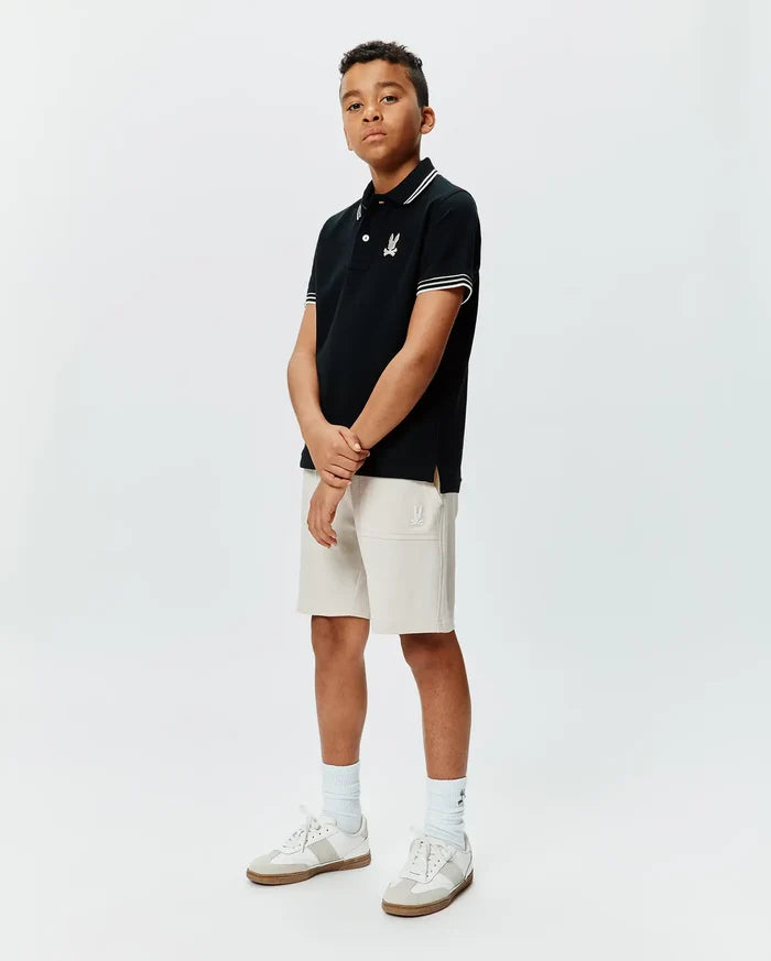 Kids Brad Pique Polo in black with white collar and sleeve tipping, featuring an embroidered Psycho Bunny skull logo on the left chest, paired with light beige knit shorts with an embroidered Psycho Bunny logo.