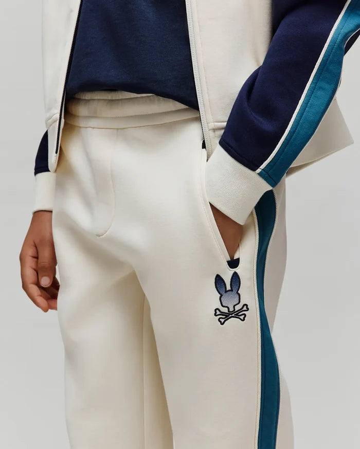 Kids Caspian Contrast Stripe Track Pant in cream french terry, featuring a relaxed fit, navy and teal side stripes with white piping, and an embroidered gradient blue Psycho Bunny skull logo on the left thigh.