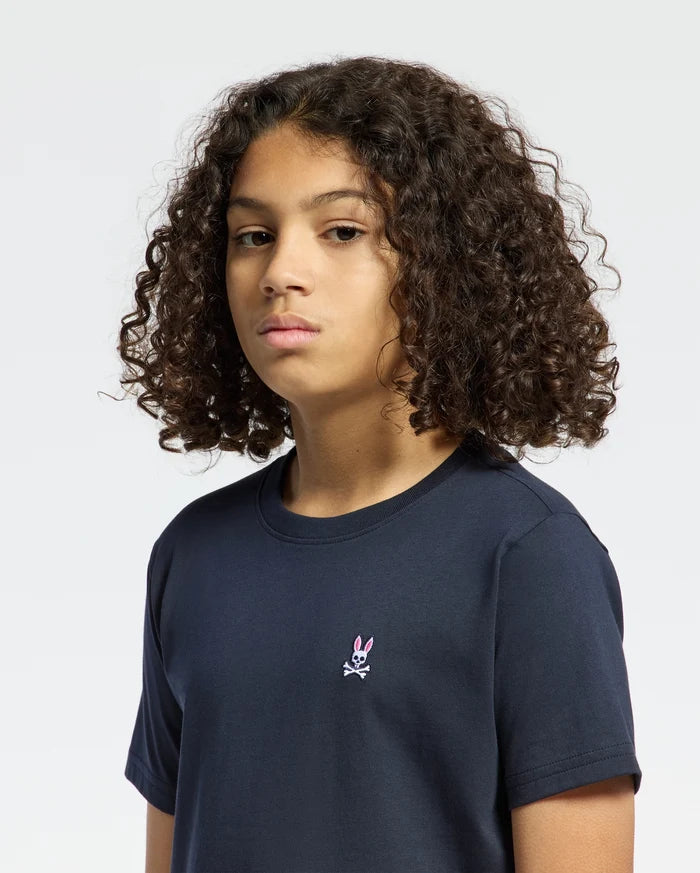 A person with curly hair wears a Psycho Bunny KIDS CLASSIC CREW NECK TEE - B0U014CRPC in navy blue, made from 100% Pima cotton, featuring a small pink bunny and crossbones logo on the chest. The plain white background complements this fun back-to-school outfit perfectly.