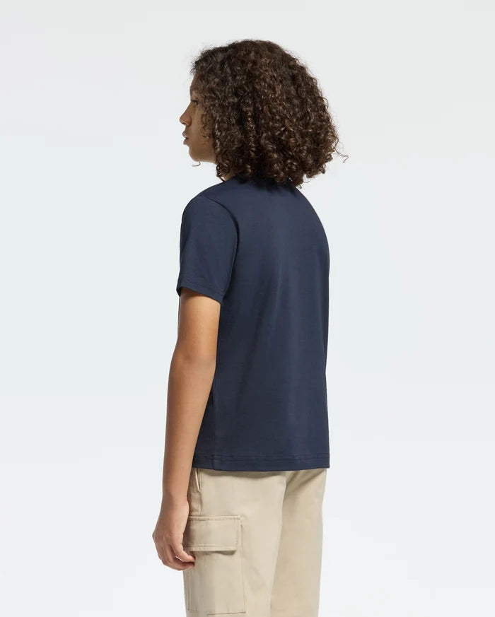A person with curly hair stands in profile, wearing Psycho Bunny's navy blue KIDS CLASSIC CREW NECK TEE - B0U014CRPC and beige cargo pants against a plain light backdrop. Their head is turned slightly, showing their side face—a perfect back-to-school look for effortless style statements.