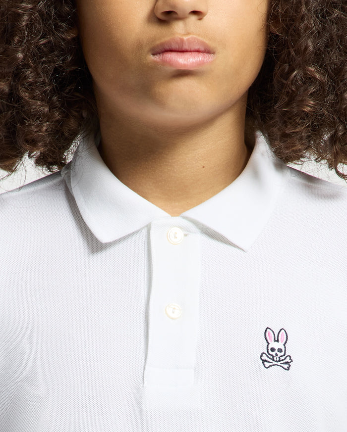 A curly-haired person wears a KIDS CLASSIC PIQUE POLO SHIRT by Psycho Bunny, ideal for back-to-school outfits. This white shirt stands out with its unique chest logo—a pink-eared rabbit above crossed bones.