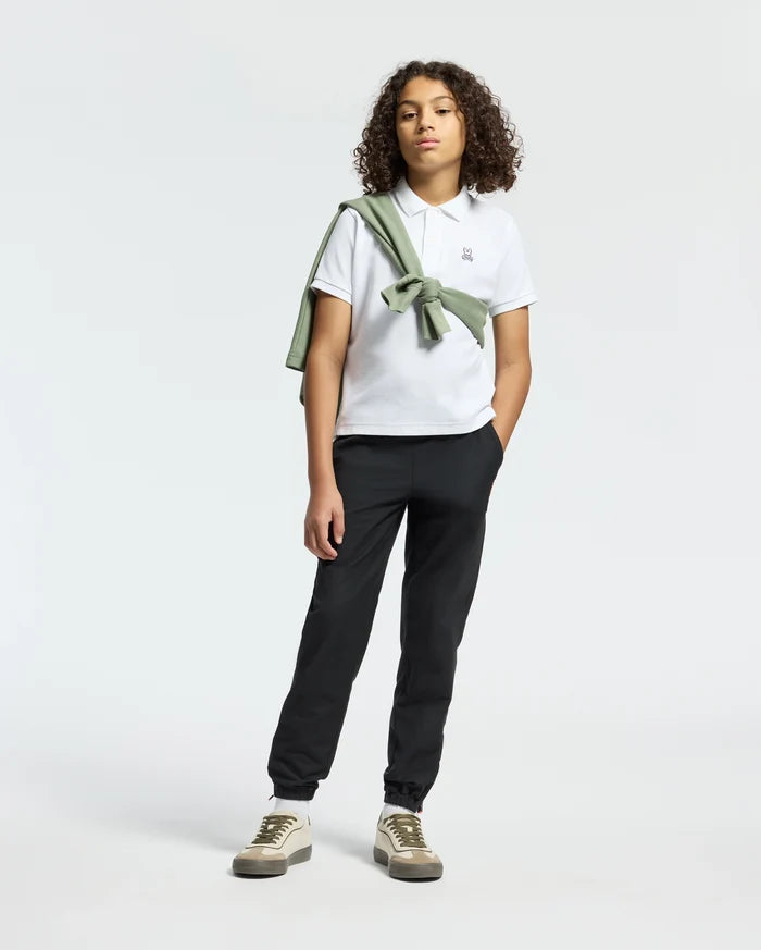 A teen confidently poses in Psycho Bunny’s KIDS CLASSIC PIQUE POLO SHIRT, paired with black pants. A green sweater drapes over their shoulder, adding back-to-school flair. With curly hair and sneakers, they stand against a neutral backdrop, ready for any occasion.