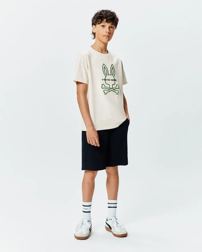 Kids Craig Graphic Tee in cream, featuring a large embroidered green Psycho Bunny skull and crossbones logo with text on the center chest, paired with black shorts. Classic fit jersey knit.