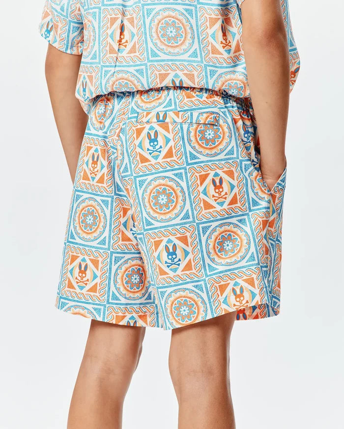Kids Frank Swim Trunk in a relaxed fit, featuring an all-over white, blue, and orange tiled geometric and floral pattern with repeated Psycho Bunny skull logos on smooth swim fabric.