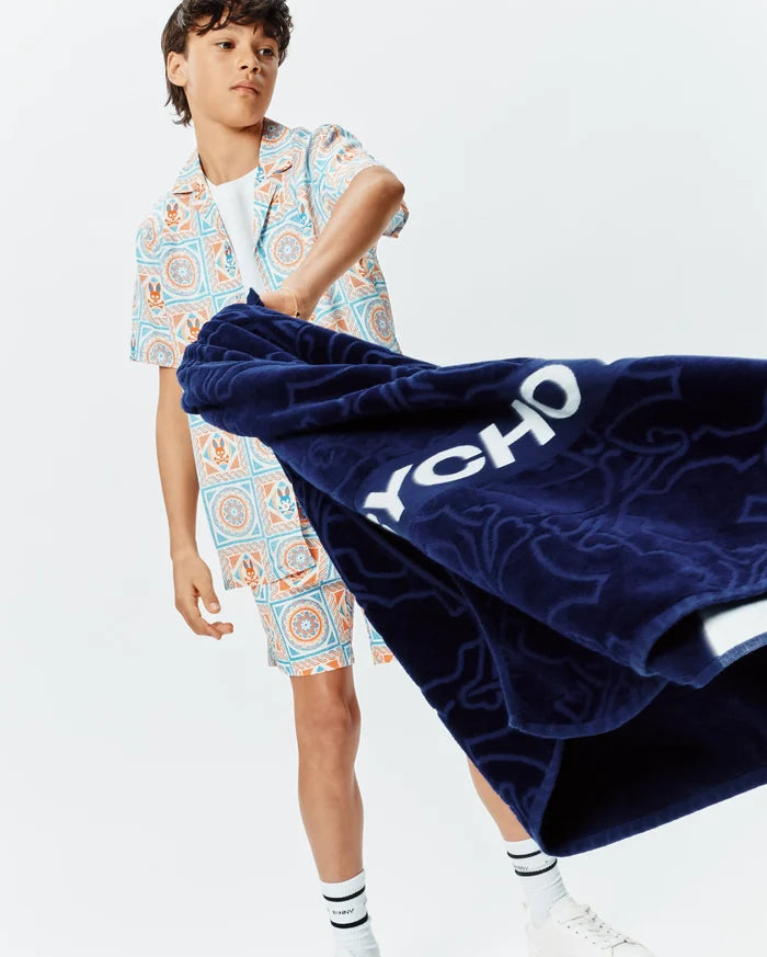 Relaxed fit short-sleeve shirt and Kids Frank Swim Trunk KB1000043 in a white, blue, orange geometric tile pattern with Psycho Bunny skull logos. Navy terry towel with white PSYCHO text and embossed bunny skulls.