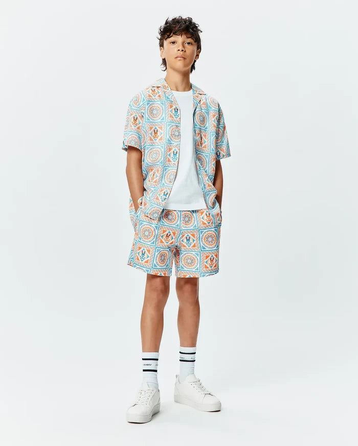 Kids Frank Swim Trunk and matching short-sleeve shirt in a relaxed fit, featuring a vibrant light blue and orange geometric tile pattern with integrated small Psycho Bunny skull logos throughout the smooth fabric.