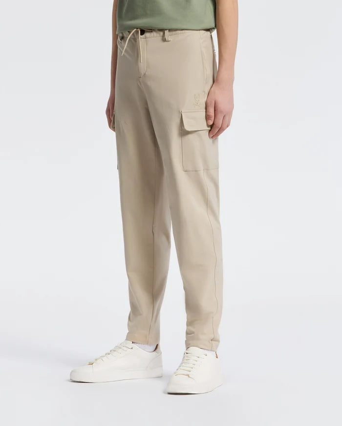 A person wears Psycho Bunny's KIDS GREEN VALLEY CARGO PANT - B0P199E200 and a green shirt against a white backdrop. The cargo pants boast pockets and a drawstring waist for comfort, paired with white sneakers.