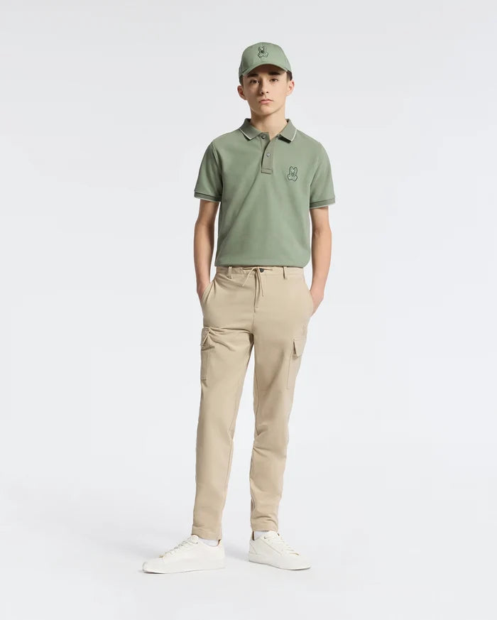 A teen wearing a green cap and matching polo shirt features the Psycho Bunny KIDS GREEN VALLEY CARGO PANT - B0P199E200, known for its cargo pockets and comfy light beige fabric, set against a white background. The outfit is completed with white sneakers and hands in pockets as he faces forward.