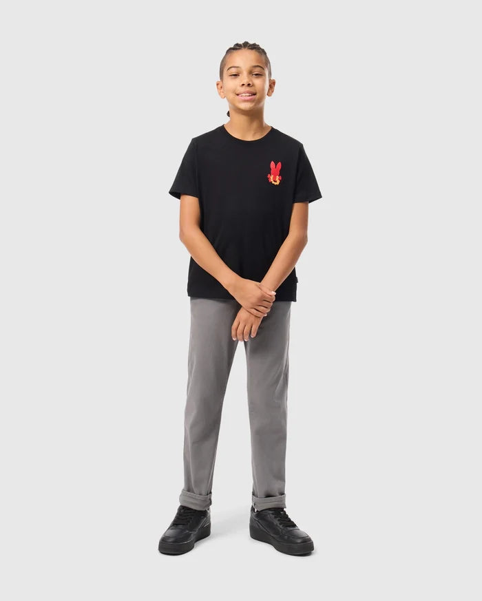A young boy stands confidently against a light gray background, wearing the KIDS HAWAII CITY TEE from Psycho Bunny, made of soft Pima cotton. His black tee features a red bunny logo on the chest and is paired with gray pants and black sneakers. With braided hair and a slight smile, he clasps his hands loosely.