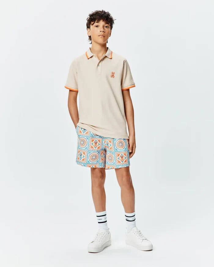 Kids Holden Pique Polo in beige with orange tipped collar and cuffs, featuring a small embroidered orange Psycho Bunny skull logo on the left chest, paired with patterned shorts.