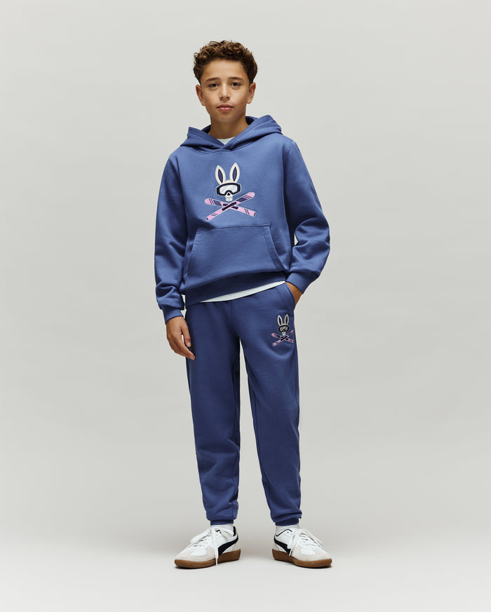 Indigo blue relaxed fit french terry hoodie and Kids Indigo Ski Bunny Sweatpant set, featuring a large chest and smaller left thigh Psycho Bunny skull logo with pink ski goggles and crossed skis.