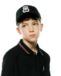 Black pique polo with red and white striped collar, small embroidered Psycho Bunny logo on chest, and a Black Kids Jaydon Baseball Cap with a large embroidered bunny skull logo.