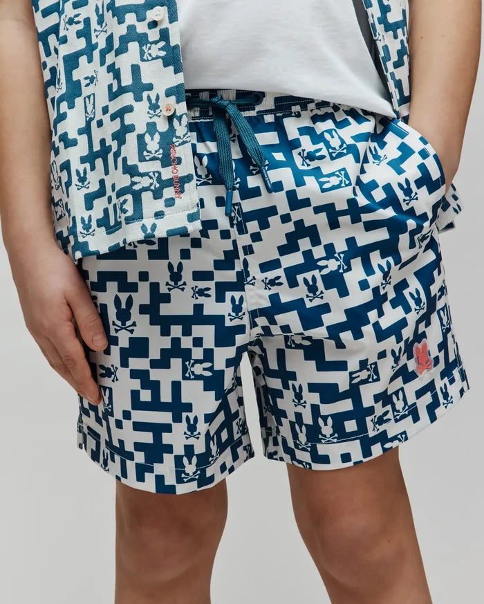 Kids Josh Swim Trunk in a relaxed fit, featuring a white and teal pixelated geometric pattern with integrated Psycho Bunny skull logos, smooth quick-dry fabric, a teal drawstring, and a small coral logo patch on the left leg.