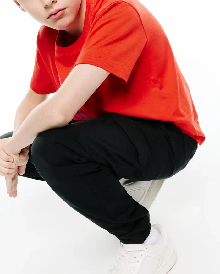 Kids Luka Cargo Pant in black, a relaxed fit streetwear style with visible side pockets and elastic cuffs, paired with a vibrant red jersey t-shirt featuring a subtle blue Psycho Bunny logo on the left chest.