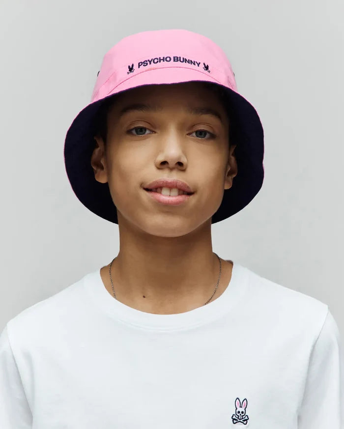 White classic fit jersey t-shirt with a small embroidered Psycho Bunny skull logo on the left chest, paired with a Kids Luke Reversible Bucket Hat in pink with a navy underside and embroidered Psycho Bunny text.