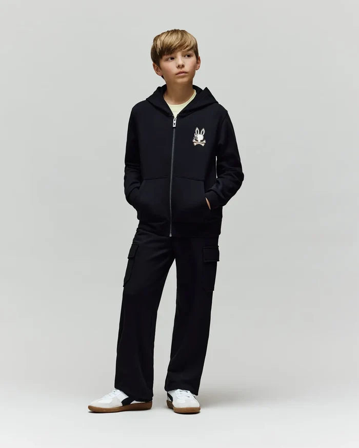 A young boy stands with hands in his pockets, wearing a black zip-up hoodie with Psycho Bunny’s cartoon bunny logo, KIDS NASH CARGO SWEATPANT (KB0800004) with cargo pockets, and white sneakers against a plain light gray background.