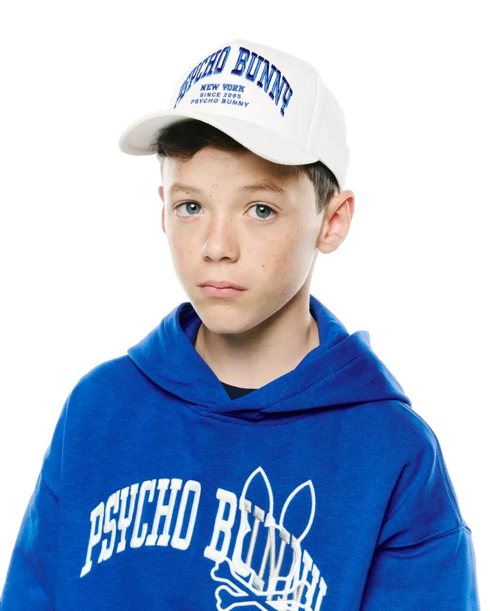 Royal blue relaxed hoodie with large white embroidered Psycho Bunny skull logo on chest, and a white Kids Nathaniel A-Frame Trucker Cap with blue embroidered Psycho Bunny New York text.