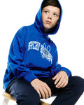 Kids Nathaniel Relaxed Hoodie in vibrant royal blue, featuring a large white Psycho Bunny text and iconic bunny skull graphic on the central chest, with a smooth jersey texture and ribbed cuffs.