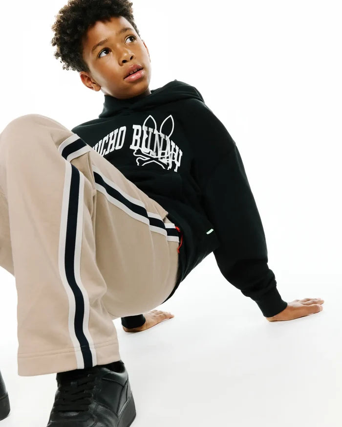 Kids Nathaniel Relaxed Hoodie in black french terry with a bold white Psycho Bunny skull logo print on the chest, complemented by relaxed beige track pants with black and white side stripes.