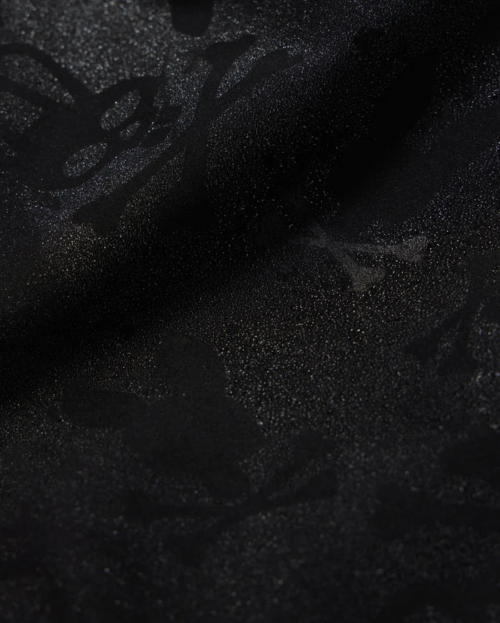 A detailed close-up of a Psycho Bunny KIDS PARKER HYDROCHROMIC SWIM TRUNK - B0W646C200 fabric displaying a subtle textured pattern with skull and crossbones and other abstract shapes. The slightly shiny, quick-dry material adds depth and dimension to the dark hydrochromatic pattern.