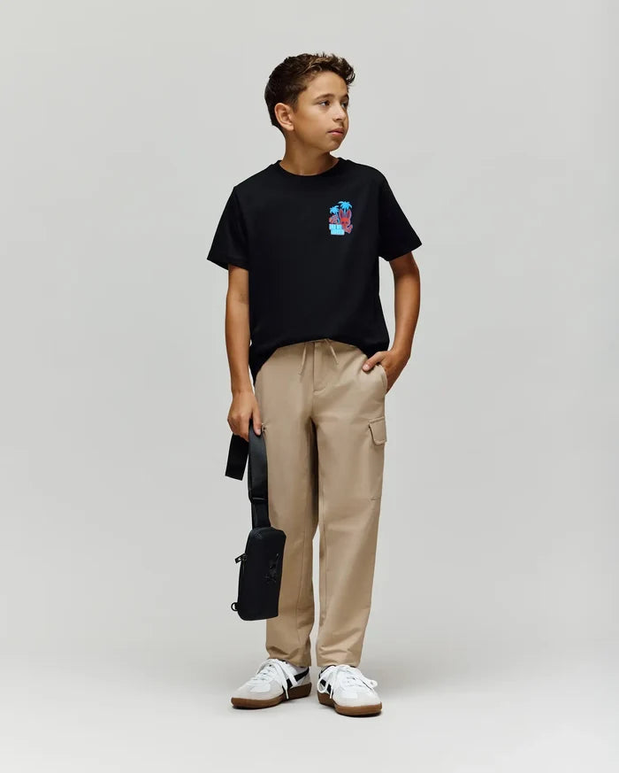 A young boy models the Psycho Bunny KIDS PUERTO RICO TEE (KT0200133), a black Pima cotton t-shirt with a colorful bunny graphic, paired with beige cargo pants, white sneakers, and a black bag, looking to his right against a plain backdrop.