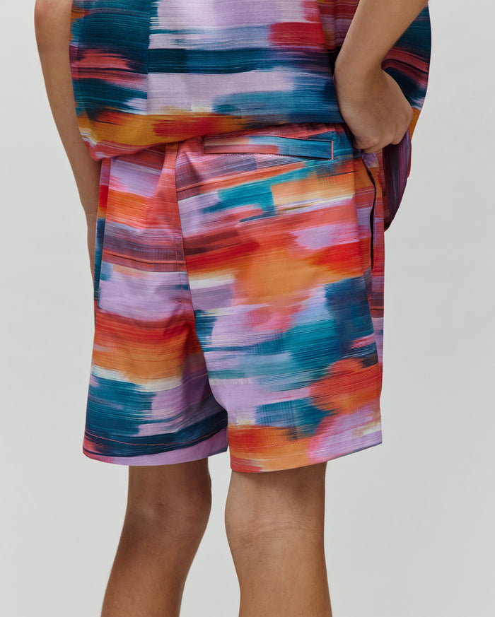 Kids Raphael Swim Trunk, a Psycho Bunny piece, showcasing a vibrant multi-color brushstroke pattern in teal, orange, red, purple, and pink, with a relaxed fit and smooth fabric.