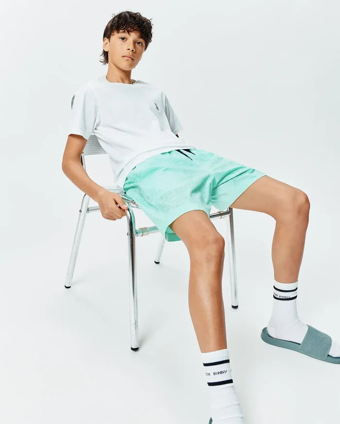 White t-shirt with small embroidered Psycho Bunny skull logo on chest, paired with Kids Samos Hydrochromic Printed Swim Trunk in mint green featuring a subtle all-over bunny skull pattern, and white Psycho Bunny socks.