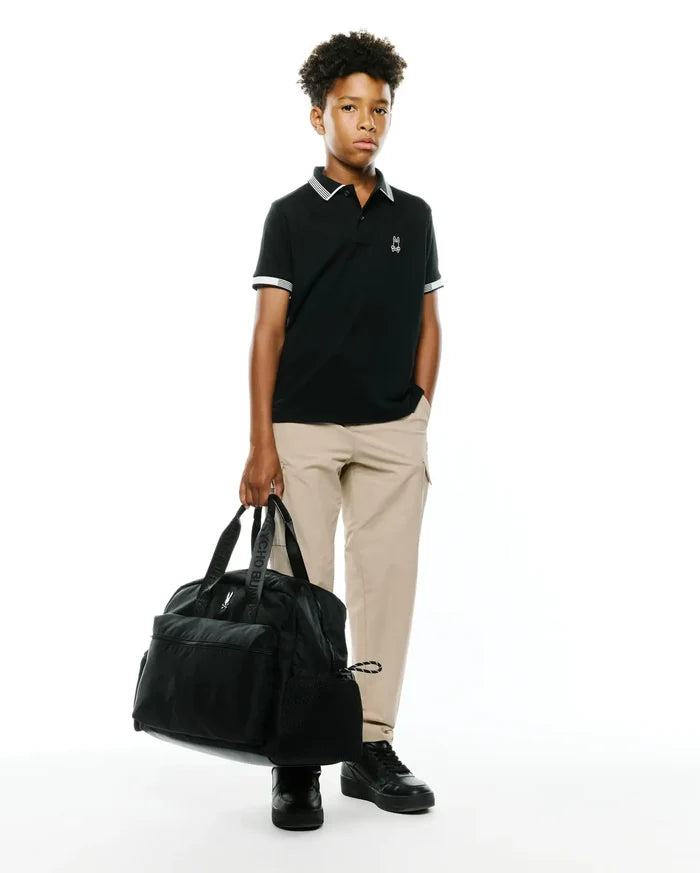 Kids Timmy Pique Polo in classic black pique with white striped collar and cuffs, featuring a small embroidered white Psycho Bunny skull logo on the left chest, styled with khaki pants.