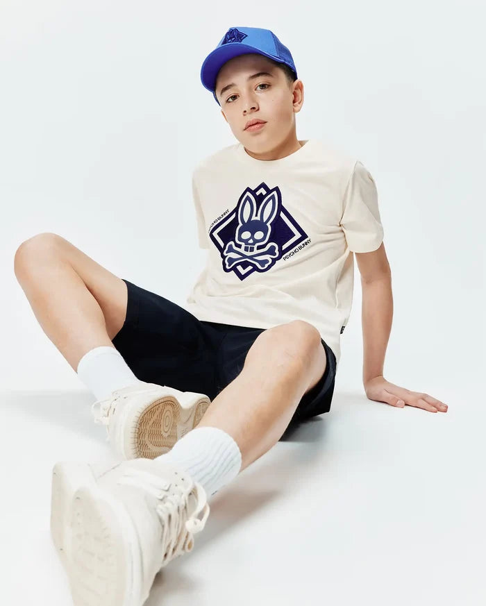 Kids Tobias Graphic Tee in cream jersey with a large navy blue Psycho Bunny skull logo on the chest, paired with navy shorts and a royal blue mesh baseball cap.