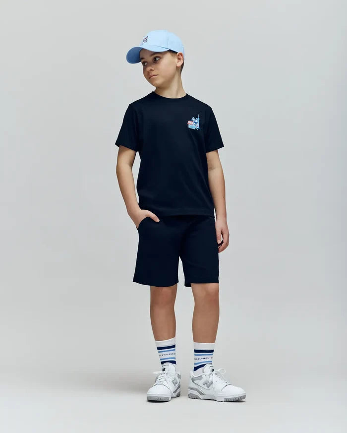 A boy in a light blue cap, black Psycho Bunny KIDS TORONTO TEE (B0U383F200), black shorts, white sneakers, and white striped socks stands with one hand in his pocket against a plain light background.